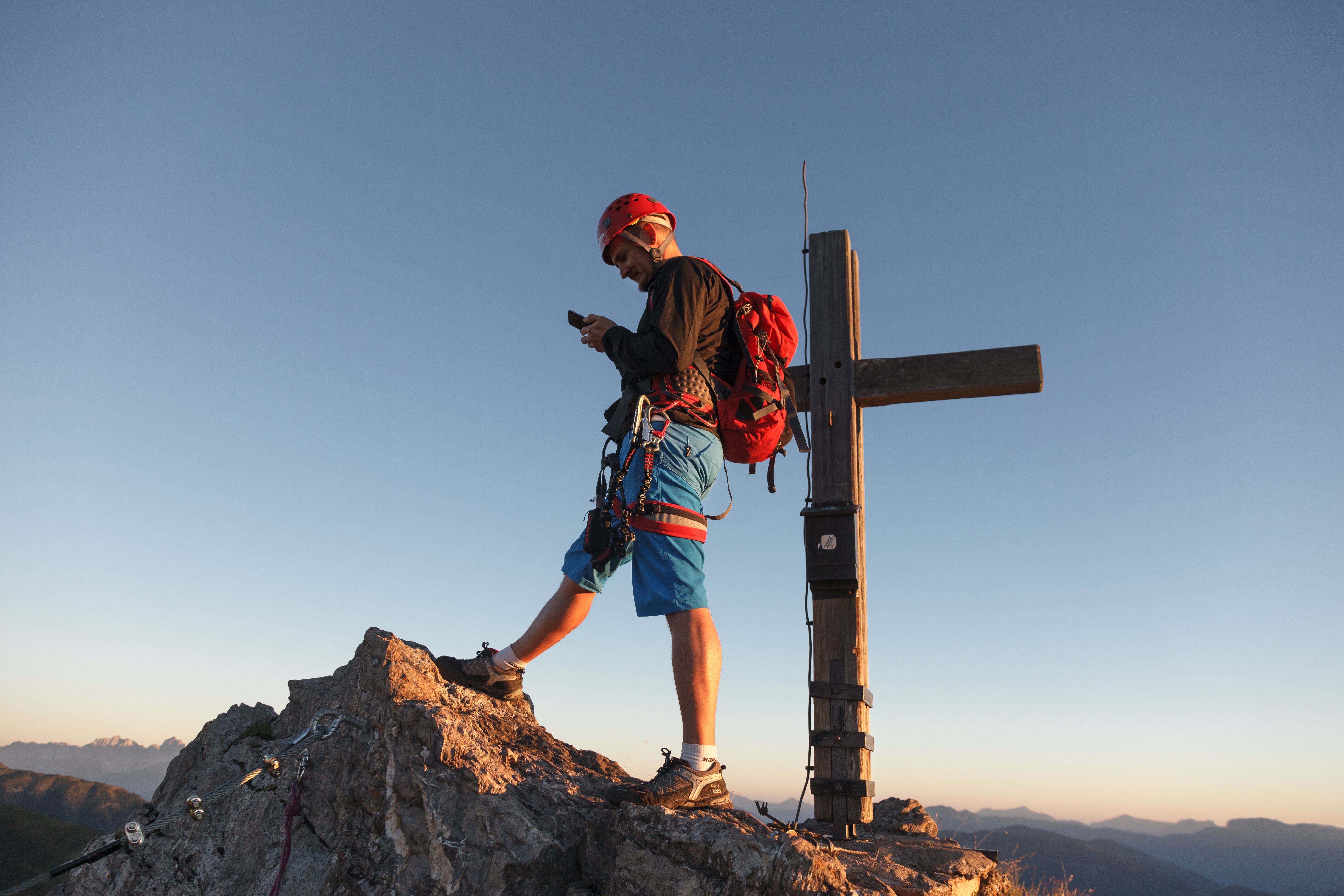 Bergsteiger mit Handy vor Gipfelkreuz